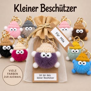 Könnte beinhalten: Eine Sammlung flauschiger, runder Pompon-Schlüsselanhänger mit gehäkelten Mützen und weißen Augen, präsentiert in einem Organzabeutel mit einem Etikett "Für Mila" und "Ich bin dein kleiner Beschützer," sowie Text "Kleiner Beschützer" und "VIELE FARBEN ZUR AUSWAHL."