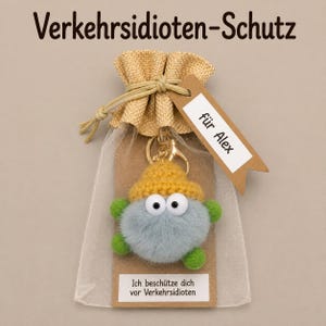 Könnte beinhalten: Ein flauschiger, runder, hellblauer Kreaturen-Schlüsselanhänger mit gelber Häkelmütze und grünen Gliedmaßen in einem transparenten Beutel. Ein braunes Etikett am Beutel trägt die Aufschrift "für Alex" und ein Schild auf der Karte "Ich beschütze dich vor Verkehrsidioten".
