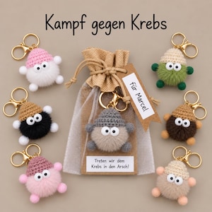 Könnte beinhalten: Sieben Plüsch-Schlüsselanhänger mit gehäkelten Mützen und Wackelaugen, angeordnet um einen Jute-Geschenkbeutel. Der Beutel hat ein Etikett mit der Aufschrift 'für Marcel' und 'Treten wir dem Krebs in den Arsch!', sowie 'Kampf gegen Krebs' darüber.