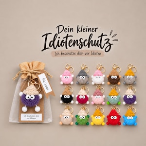 Könnte beinhalten: Eine Sammlung von fünfzehn kleinen, runden, gehäkelten Pompon-Schlüsselanhängern mit spitzen Mützen und Wackelaugen, in einem Raster angeordnet. Ein Anhänger befindet sich in einem Jutebeutel mit einem Etikett "Für Lena" und "Ich beschütze dich vor Idioten."