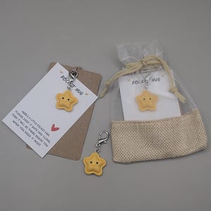 Könnte beinhalten: Ein Pocket Hug Geschenkset mit einem gelben Sternanhänger mit einem lächelnden Gesicht. Das Set enthält einen Schlüsselanhänger, eine Karte mit Text und einen kleinen Jutebeutel. Auf der Karte steht "Pocket Hug".