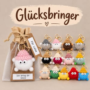 Könnte beinhalten: Eine Sammlung kleiner, runder, gehäkelter Plüsch-Schlüsselanhänger mit Wackelaugen und spitzen Mützen in verschiedenen Farben. Ein Anhänger befindet sich in einem kleinen Jute-Geschenkbeutel mit einem Etikett mit der Aufschrift Für Luisa und Ich bring dir Glück.
