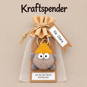Könnte beinhalten: Ein Schlüsselanhänger mit einer flauschigen beigen Pompon-Figur, einer gelben Häkelmütze und großen weißen Augen, befestigt an einer braunen Karte in einem Organzabeutel. Die Karte trägt den Text "Ich bin dein kleiner Kraftspender".