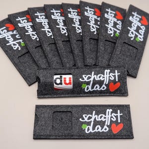 Könnte beinhalten: Eine Sammlung dunkelgrauer Filz-Getränkehalter mit dem Schriftzug "schaffst das" in weißer Schrift, einem roten Herz und einem grünen Kleeblatt. Ein Halter hält ein kleines weiß-rotes Objekt. Die Halter sind fächerförmig angeordnet.