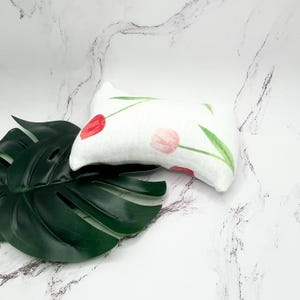 May include: A small, white rectangular pillow with a floral tulip design in red and pink. Green stems and leaves accent the flowers. The pillow is placed on a large, dark green leaf.
