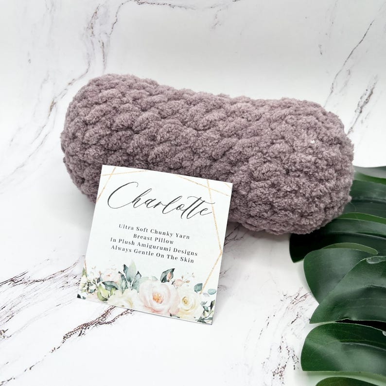 May include: A mauve-colored, chunky yarn breast pillow with a white tag that reads "Charlotte Ultra Soft Chunky Yarn Breast Pillow In Plush Amigurumi Designs Always Gentle On The Skin".