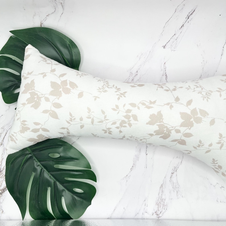 Może przedstawiać: Długa, prostokątna poduszka typu body pillow z białym materiałem w powtarzający się wz&oacute;r jasnobeżowych kwiat&oacute;w i pnączy. Poduszka jest ułożona ukośnie w kadrze, otoczona dwoma dużymi, ciemnozielonymi liśćmi monstery o tropikalnym wyglądzie.