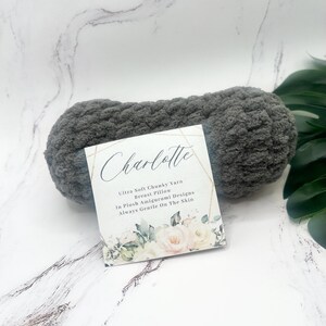 May include: A gray yarn ball with a white label that says "Charlotte Ultra Soft Chunky Yarn Breast Pillow In Plush Amigurumi Designs Always Gentle On The Skin". The label has a floral design.