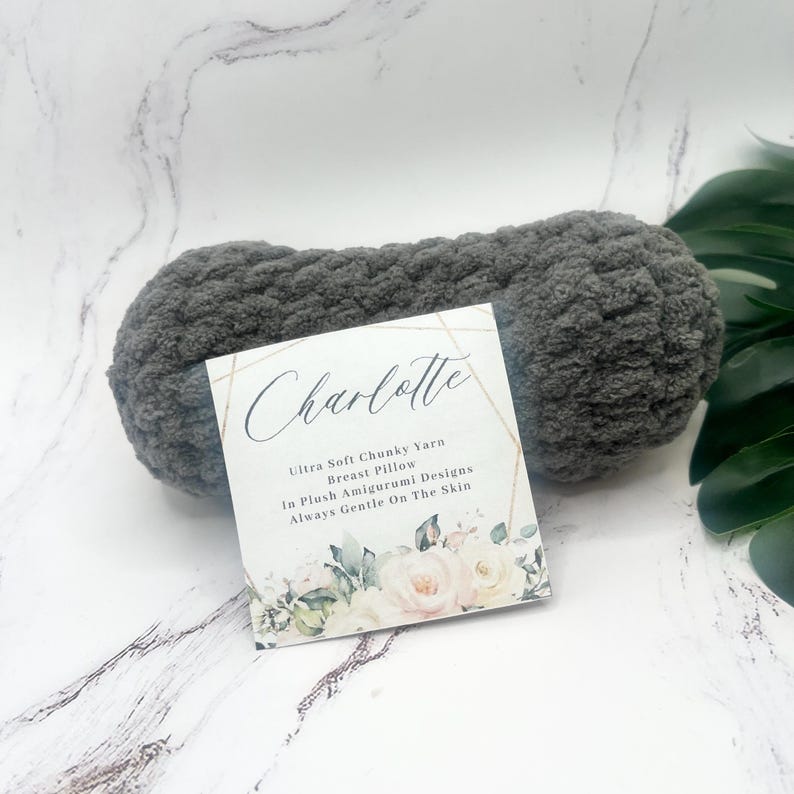 May include: A gray yarn ball with a white label that says "Charlotte Ultra Soft Chunky Yarn Breast Pillow In Plush Amigurumi Designs Always Gentle On The Skin". The label has a floral design.