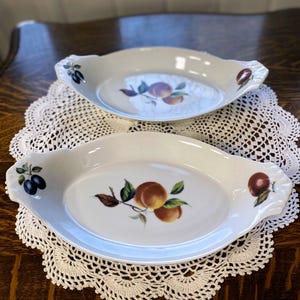 May include: Two white oval serving dishes with handles, each featuring a fruit design. One dish has plums, the other peaches, with green leaves. The dishes rest on a white lace doily on a wooden table.