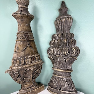 May include: Two decorative finials with a weathered, distressed finish in shades of brown, gray, and gold. The left finial features a pineapple-shaped top, while the right finial displays a tiered, scroll-like design with leaf-shaped accents.