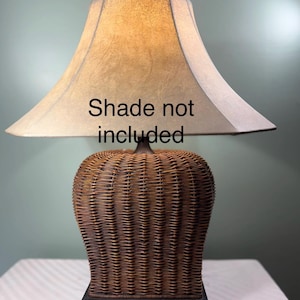 May include: A table lamp with a brown woven wicker base and a square black pedestal. The lamp features a bell-shaped beige lampshade with the text Shade not included printed in black across the center of the shade surface.