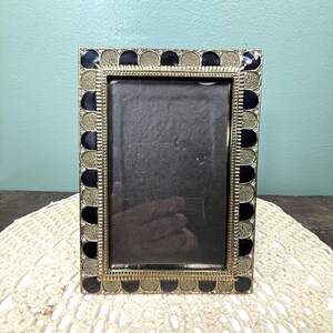 May include: A rectangular picture frame featuring a metallic gold finish with a textured, scalloped border pattern. The frame is accented with alternating black enamel inlays and small, clear embedded stones along the outer edge.