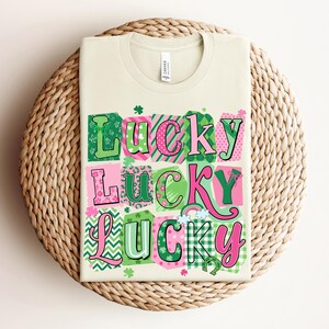 Pode incluir: Uma camiseta creme com a palavra "Lucky" em um design de patchwork colorido. A palavra está em rosa e verde, com trevos e um arco-íris. A camiseta está dobrada em um tapete tecido.