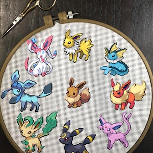 Puede incluir: Un aro de bordado de punto de cruz con una variedad de personajes de Pokémon, incluyendo Eevee, Vaporeon, Jolteon, Flareon, Espeon, Umbreon, Leafeon, Glaceon y Sylveon.