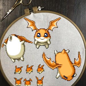 May include: A cross-stitch project featuring pixelated orange and white characters from a cartoon series. The embroidery is in a wooden hoop, with gold scissors resting on the dark surface.