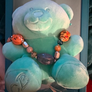 May include: A light teal plush teddy bear wearing a beaded necklace with orange and gray beads. The bear has embroidered initials on its paw and is displayed in a teal frame.