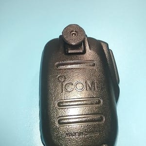 May include: A black ICOM handheld microphone with a textured surface. The microphone has the brand name "ICOM" printed on the front and the words "MADE IN" are visible. The microphone is set against a light blue background.