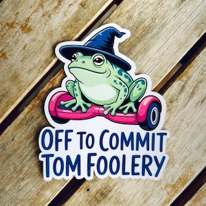Könnte beinhalten: Aufkleber mit einem Cartoon-Frosch mit blauem Zaubererhut, der auf einem rosa Hoverboard fährt. Der Text "OFF TO COMMIT TOM FOOLERY" steht darunter. Auf einer Holzoberfläche.