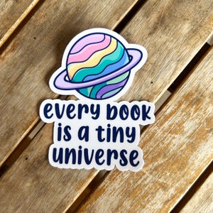 Könnte beinhalten: Ein weißer Aufkleber mit einem farbenfrohen Planeten-Design und dem Text "every book is a tiny universe". Der Planet hat ein violettes, blaues, gelbes und rosa Farbschema mit einem violetten Ring. Der Aufkleber befindet sich auf einer Holzoberfläche.