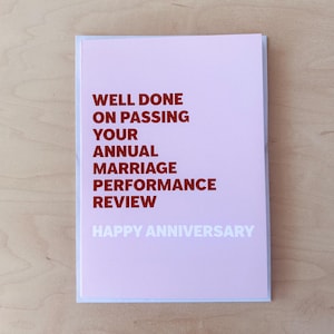 May include: A pink greeting card with the text "WELL DONE ON PASSING YOUR ANNUAL MARRIAGE PERFORMANCE REVIEW" in red and "HAPPY ANNIVERSARY" in white. The card is rectangular and has a white border.