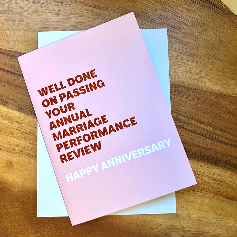May include: A pink greeting card with the text "WELL DONE ON PASSING YOUR ANNUAL MARRIAGE PERFORMANCE REVIEW HAPPY ANNIVERSARY" in white and red lettering. The card is on top of a light blue card, set against a wooden surface.