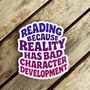 May include: A white sticker with a purple and pink gradient border and text that reads "READING BECAUSE REALITY HAS BAD CHARACTER DEVELOPMENT" in a retro font. The sticker is on a wooden surface.