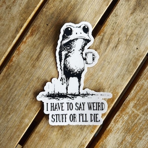 May include: A black and white sticker featuring a cartoon frog holding a mug. The frog has large eyes and a slightly sad expression. The sticker has the text: "I HAVE TO SAY WEIRD STUFF OR I'LL DIE."
