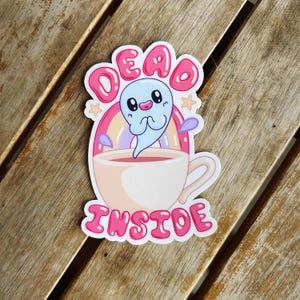 May include: A sticker featuring a cartoon seal in a teacup with the words "DEAD INSIDE" in pink bubble letters. The seal is light blue with a sad expression, and the teacup is filled with a pink liquid.