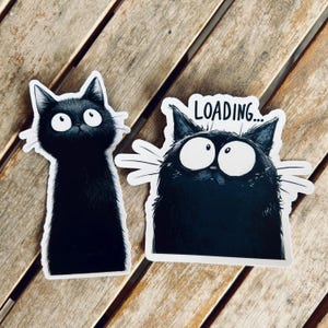 May include: Two black cat stickers with white outlines. One cat is tall and slender, the other is rounder with the word "LOADING..." above it. Both cats have large, expressive eyes and white whiskers, set against a wooden background.