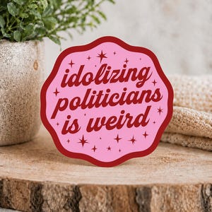 May include: A scalloped-edge sticker with a light pink background and a dark red border. The text "idolizing politicians is weird" is printed in dark red cursive lettering, surrounded by small dark red star icons scattered across the surface.