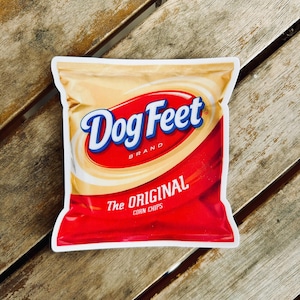 May include: A bag of Dog Feet brand corn chips. The bag is red and tan with the words "Dog Feet" in blue script. The text "The Original Corn Chips" is also visible.