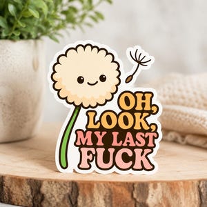 Op de afbeelding: Een sticker met een cartoon paardenbloem met een lachend gezicht. De tekst "OH, LOOK, MY LAST FUCK" staat in oranje en roze. De sticker is geplaatst op een houten oppervlak.