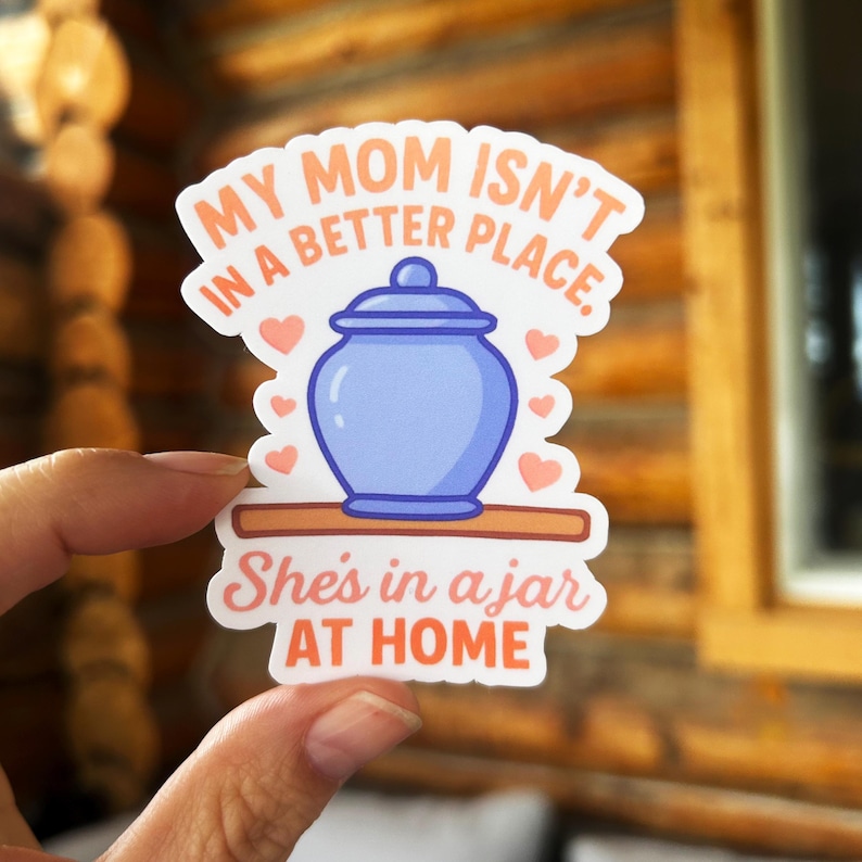 May include: A sticker with the text "MY MOM ISN'T IN A BETTER PLACE. She's in a jar AT HOME." The sticker features a blue jar with a lid, surrounded by pink hearts, on a wooden shelf.