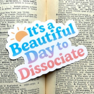 May include: A die-cut sticker with the phrase "It's a Beautiful Day to Dissociate" in blue, purple, and pink, with a yellow and orange sun graphic. The sticker is on a background of old book pages.