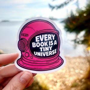 Può includere: Un adesivo rosa con un design a casco da astronauta e la scritta bianca "EVERY BOOK IS A TINY UNIVERSE". L'adesivo ha un contorno nero ed è mostrato su uno sfondo sfocato all'aperto.