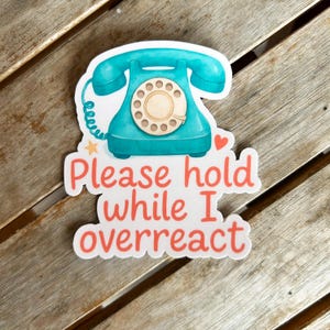 Può includere: Un adesivo con l'illustrazione di un telefono a disco turchese. L'adesivo presenta il testo "Please hold while I overreact" in rosso e bianco. L'adesivo è su una superficie di legno.