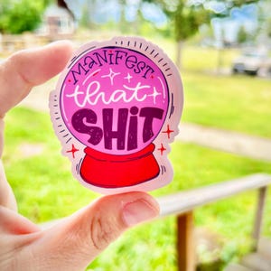 May include: A pink sticker with the words "Manifest that SHIT" in a stylized font, set within a crystal ball design. The sticker has a red base and is held against a blurred green background.