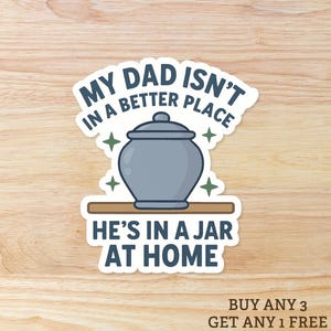 May include: A sticker with the text "MY DAD ISN'T IN A BETTER PLACE HE'S IN A JAR AT HOME". The sticker features a gray jar with a lid, sitting on a brown shelf, with green star accents.