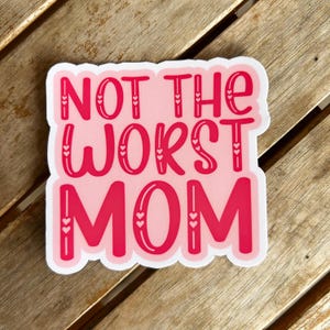 May include: A pink and red sticker with the text "NOT THE WORST MOM". The sticker has a white border and heart accents. The sticker is on a wooden surface.