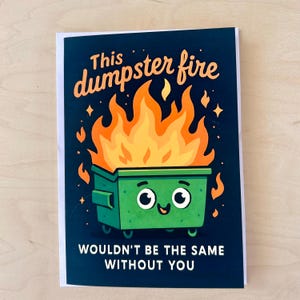 May include: A greeting card with a navy blue background and a cartoon illustration of a green dumpster on fire. The text reads "This dumpster fire wouldn't be the same without you" in orange and white.