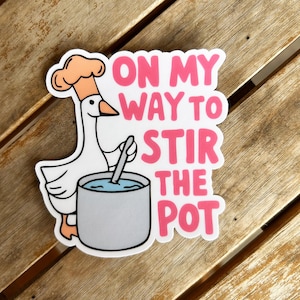 May include: A white goose wearing a chef's hat and holding a spoon, stirring a pot of blue liquid. The sticker has the text "ON MY WAY TO STIR THE POT" in pink letters with a white outline. The sticker is on a wooden surface.