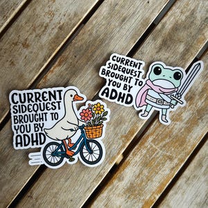 May include: Two stickers with white borders on a wooden surface. One sticker features a goose riding a blue bicycle with a basket of flowers, and the text "CURRENT SIDEQUEST BROUGHT TO YOU BY ADHD." The other sticker shows a frog with a sword and the same text.