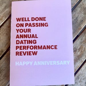 May include: A pink greeting card with the text "WELL DONE ON PASSING YOUR ANNUAL DATING PERFORMANCE REVIEW" in red and "HAPPY ANNIVERSARY" in white. The card is on a wooden surface.
