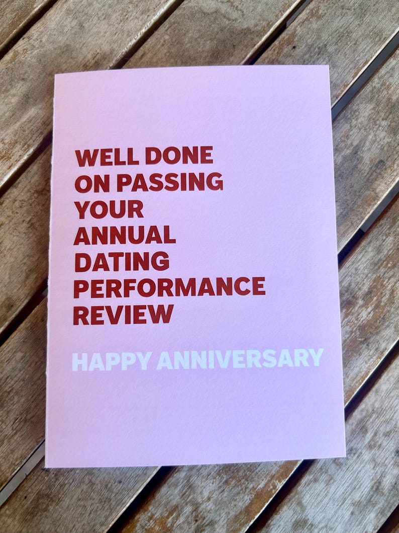 May include: A pink greeting card with the text "WELL DONE ON PASSING YOUR ANNUAL DATING PERFORMANCE REVIEW" in red and "HAPPY ANNIVERSARY" in white. The card is on a wooden surface.