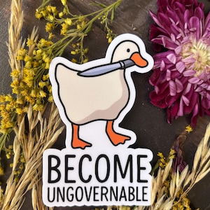 Könnte beinhalten: Weißer Aufkleber mit einer Comic-Gans, die ein Messer im Schnabel hält. Der Aufkleber trägt die schwarzen Worte "BECOME UNGOVERNABLE". Umgeben von gelben Blumen und einem dunklen Hintergrund.