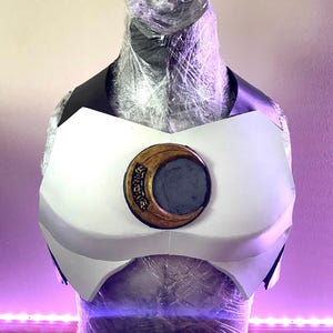 Puede incluir: Una armadura de pecho blanca con un emblema dorado y gris. La armadura tiene detalles en negro y se exhibe sobre una forma cubierta con envoltura de plástico. El emblema presenta un diseño de media luna con texto.