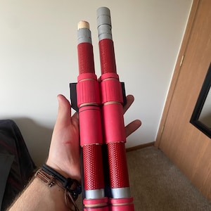 May include: Two red and gray foam-covered prop weapons with a black handle. The weapons are held in a person's hand.