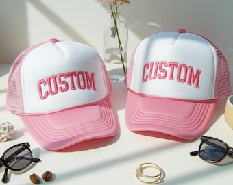 Könnte beinhalten: Zwei rosa-weiße Trucker-Caps mit dem Wort "CUSTOM" in Rosa gestickt. Die Caps haben einen rosa Schirm und eine weiße Frontplatte. Das Mesh-Rückenteil ist rosa. Sonnenbrillen und ein goldener Ring sind ebenfalls sichtbar.