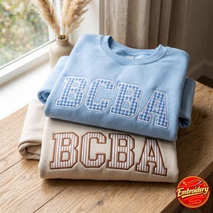May include: A light blue crewneck sweatshirt featuring the letters BCBA in a blue and white gingham pattern, layered over a beige sweatshirt with the letters BCBA in a brown and white vertical striped pattern, both with white embroidery outlines.